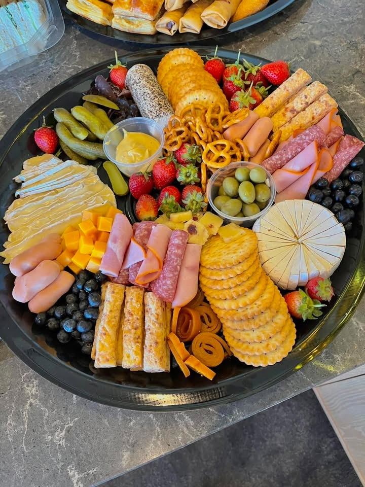 Round cheese platter with a brie wheel, cheddar cubes, cold meats, crackers, pretzels, olives, gherkins, strawberries and blueberries